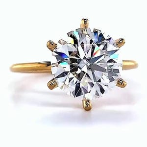 May include: A gold ring with a large, round, clear gemstone. The gemstone is held in place by six prongs. The ring band is a simple, thin gold band. The brand name "VEVA JEWEL" is printed at the bottom of the image.