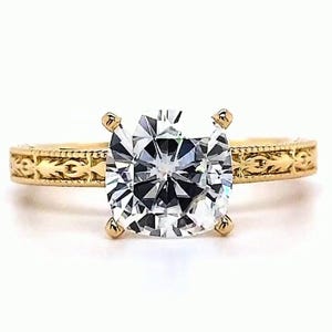 May include: A gold engagement ring with a large, clear, square-cut diamond. The band features an engraved pattern. The ring is set against a white background. The brand name "VEVA JEWEL" is at the bottom.