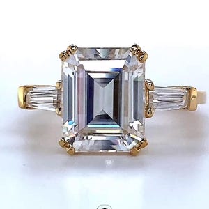 May include: A gold engagement ring with an emerald-cut center stone and two baguette side stones. The ring features a four-prong setting and is set against a white background. The ring is a classic and elegant design.