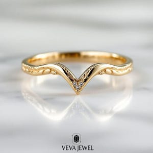 May include: A gold ring with a V-shaped design. The ring has a detailed leaf pattern on the band and small, clear gemstones at the point of the V. The ring is shown on a reflective surface. The brand name "VEVA JEWEL" is at the bottom.