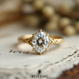 May include: A gold engagement ring with an oval-cut diamond. The ring features an ornate, textured band and a detailed setting around the diamond. The ring is set on a white surface with the text "VEVA JEWEL".