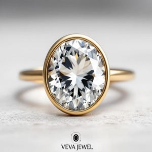 May include: A gold ring with a large, oval-cut diamond. The diamond is set in a bezel setting, and the band is a simple, polished gold. The ring is on a white surface. The text "VEVA JEWEL" is at the bottom.