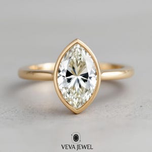 May include: A marquise-cut diamond ring with a gold band. The diamond is set in a bezel setting, and the ring is displayed on a neutral background. The ring is from the brand VEVA JEWEL.