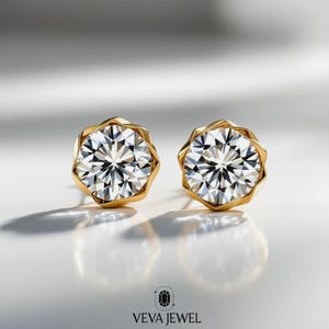 May include: A pair of gold stud earrings with a large, round, clear gemstone. The gemstones are faceted, reflecting light. The earrings have a unique, geometric gold setting. The brand name "VEVA JEWEL" is at the bottom of the image.