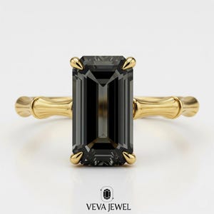 May include: A gold ring featuring a large, rectangular, dark gray gemstone. The ring has a bamboo-style band and four prongs securing the stone. The brand name "VEVA JEWEL" is at the bottom.