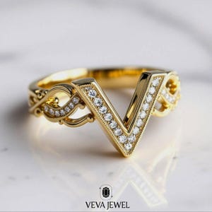 May include: A gold-colored ring with a V-shaped design, encrusted with small, sparkling stones. The ring features intricate filigree detailing on the sides and is set against a soft, neutral background. The brand name "VEVA JEWEL" is visible at the bottom.