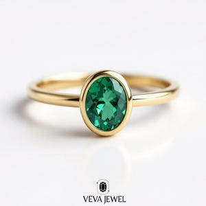 May include: A gold ring featuring a vibrant green oval-cut gemstone. The ring has a simple, elegant design with a polished gold band and a bezel setting that highlights the emerald. The ring is set against a white background.