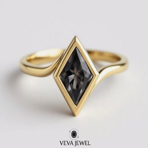 May include: A gold ring featuring a large, dark gray, diamond-shaped gemstone. The gemstone is set within a gold bezel, and the band has a smooth, curved design. The ring is displayed on a white background. The text "VEVA JEWEL" is at the bottom.
