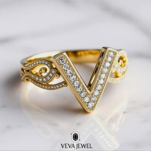 Pode incluir: Um anel dourado com um design em forma de V, cravejado com pequenos diamantes brilhantes. O anel apresenta detalhes intrincados de filigrana ao longo da banda e ao redor da forma em V, criando uma aparência elegante e luxuosa. O anel é uma joia.