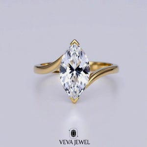 May include: A gold engagement ring with a marquise-cut diamond. The diamond is set in a prong setting and the band has a swirling design. The ring is set against a white background. The text "VEVA JEWEL" is at the bottom.
