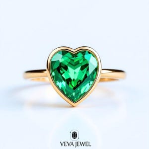 May include: A gold ring featuring a heart-shaped, faceted green gemstone. The gemstone is set in a gold bezel, and the band is a simple, polished gold. The ring is displayed on a white background. The brand name "VEVA JEWEL" is at the bottom.