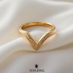 May include: A gold-colored chevron ring with an engraved geometric pattern. The ring has a V-shape design and is set against a white, silky background. The brand name "VEVA JEWEL" is at the bottom of the image.