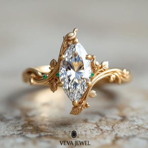 May include: A marquise-cut diamond engagement ring with a gold band. The ring features a nature-inspired design with leaf and vine details, and small green gemstones. The ring is from Veva Jewel.