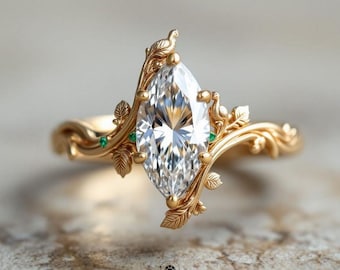 May include: A marquise-cut diamond engagement ring with a gold band. The ring features a nature-inspired design with leaf and bird details, and small green gemstones. The ring is from Veva Jewel.