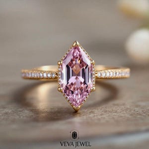 May include: A gold ring featuring a large, pink, emerald-cut gemstone. The gemstone is surrounded by a halo of smaller diamonds, and the band is also adorned with small diamonds. The ring is set against a neutral background. The text "VEVA JEWEL" is at the bottom.