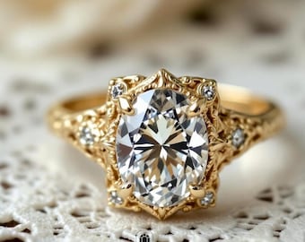 May include: A gold engagement ring with an oval-cut diamond center stone. The ring features a detailed filigree design and smaller accent diamonds. The ring is set on a white lace surface. The brand logo "VEVA JEWEL" is visible.