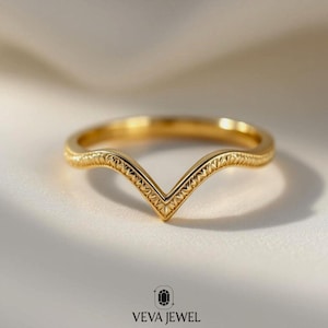 May include: A gold-colored ring with a V-shaped design. The ring has a delicate, patterned texture along the band and within the V. The ring is set against a soft, neutral background. The brand name "VEVA JEWEL" is at the bottom.