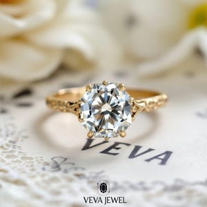 May include: A gold engagement ring with a large, clear, round-cut gemstone. The ring has an ornate, engraved band and is set against a soft, blurred background. The ring is from VEVA JEWEL.