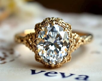 Puede incluir: Un anillo de oro con un diamante de talla ovalada. El diamante está engastado en un engaste ornamentado de filigrana. El anillo está sobre una superficie blanca con la palabra "Veva" impresa en azul.