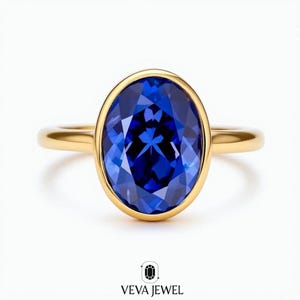 May include: A gold ring featuring a large, oval-cut, deep blue gemstone. The gemstone is set in a gold bezel, and the band is a simple, polished gold. The ring is displayed on a white background. The text "VEVA JEWEL" is at the bottom.