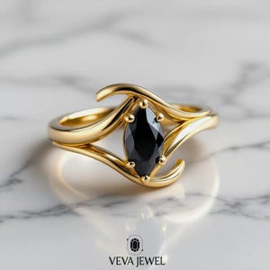 May include: A gold ring featuring a marquise-cut black gemstone. The ring has a unique, flowing design with a split shank and decorative accents. The ring is set against a marble background. The brand name "VEVA JEWEL" is at the bottom.