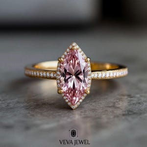 May include: A gold ring featuring a marquise-cut pink gemstone with a halo of small diamonds. The band is also adorned with small diamonds. The ring is set against a gray background. The logo "VEVA JEWEL" is at the bottom.