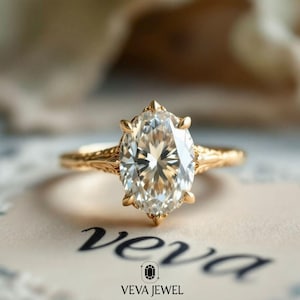 May include: A gold engagement ring with an oval-cut diamond. The ring has a detailed, textured band and is set against a soft background. The ring is from Veva Jewel.