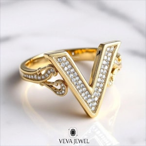 Könnte beinhalten: Ein goldfarbener Ring mit einem großen V-förmigen Design, das mit kleinen, klaren Edelsteinen besetzt ist. Der Ring hat ein dekoratives Band mit zusätzlichen Edelsteinen und verzierten Filigran-Details. Der Ring liegt auf einer weißen Oberfläche.