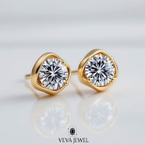 May include: A pair of gold stud earrings with a round, brilliant-cut diamond center stone. The diamonds are set in a gold bezel setting. The earrings are displayed on a white surface. The brand logo "VEVA JEWEL" is at the bottom.