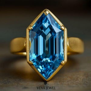 May include: A gold ring featuring a large, faceted, light blue gemstone in a unique geometric shape. The gemstone is set in a gold bezel, and the band is a simple, polished gold. The ring is displayed on a neutral surface. The text "VEVA JEWEL" is at the bottom.