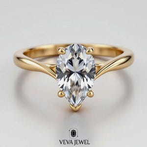 May include: A gold engagement ring with a pear-shaped diamond. The ring has a smooth, polished band and four prongs securing the large, clear gemstone. The ring is set against a white background. The text "VEVA JEWEL" is at the bottom.