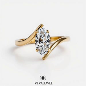 May include: A gold engagement ring with a large, oval-cut diamond. The band has a unique, flowing design that curves around the diamond. The ring is set against a white background. The text "VEVA JEWEL" is at the bottom.