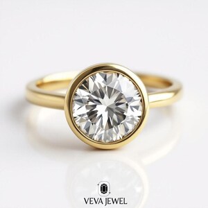 May include: A gold bezel-set engagement ring with a large, round, brilliant-cut diamond. The ring's band is a simple, polished gold. The diamond sparkles brightly against a white background. The Veva Jewel logo is at the bottom.