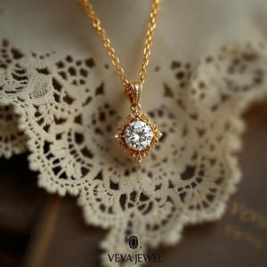 May include: A gold-colored pendant necklace featuring a large, round, clear gemstone set in an ornate, square-shaped frame. The pendant hangs from a delicate chain. The necklace is displayed on a lace surface. The brand name "VEVA JEWEL" is at the bottom.