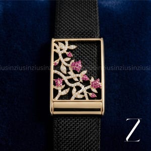 May include: A gold-coloured watch buckle with a floral design. The rectangular buckle has a black background with pink flowers and clear gemstones. The watch strap is black. The design is intricate and detailed.