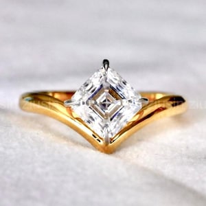 Könnte beinhalten: Ein goldener Ring mit einem großen, quadratisch geschliffenen, klaren Edelstein. Das Band hat ein V-förmiges Design. Der Ring ist vor einem weißen Hintergrund platziert. Der Buchstabe "Z" befindet sich in der unteren rechten Ecke.