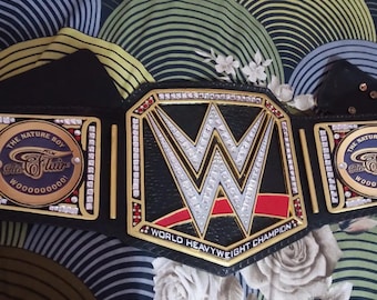 Könnte beinhalten: Ein schwarzer und goldener World Heavyweight Championship-Gürtel mit dem WWE-Logo in der Mitte. Der Gürtel trägt den Text "World Heavyweight Champion" und "The Nature Boy Ric Flair".