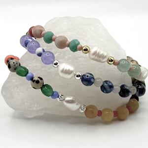 May include: Three beaded bracelets with a variety of colorful beads, including pearls, gemstones, and speckled beads. The bracelets are arranged in a layered style, showcasing a mix of colors like green, blue, pink, and yellow. The bracelets are resting on a white, textured surface.