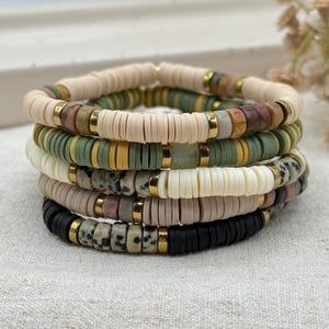 May include: Stack of five beaded bracelets in neutral tones. The bracelets feature flat, disc-shaped beads in colors like cream, olive green, black, and speckled gray. Gold-colored spacers add a touch of shine to the bracelets.
