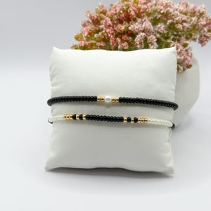 May include: Two beaded bracelets. The top bracelet is black with gold accents and a single pearl. The bottom bracelet is white with black and gold accents. The bracelets are displayed on a white pillow.