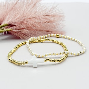 May include: Three beaded bracelets. Two bracelets feature gold-colored beads and white beads. The third bracelet has gold beads and a white cross charm. The bracelets are on a white surface with pink decorative foliage.
