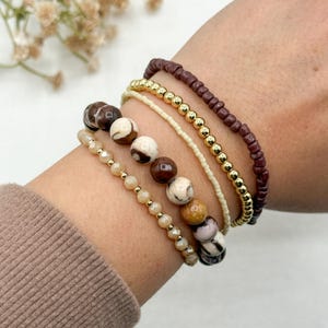 May include: A collection of beaded bracelets in various styles and colors. The bracelets include brown, gold, and cream-colored beads, as well as a dark red beaded bracelet. The bracelets are stacked on top of each other.