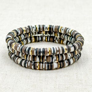 May include: Three stacked bracelets with a mix of brown, black, white, and gold-colored disc beads. The bracelets are made of small, flat, round beads strung together, creating a layered effect. The gold accents add a touch of elegance.
