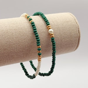 May include: Two beaded bracelets. One bracelet features dark green beads with gold accents and a single pearl. The other bracelet has dark green and white beads with gold accents. Both bracelets are displayed on a tan fabric roll.