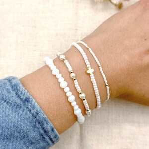May include: Four white beaded bracelets with gold accents are displayed on a wrist. One bracelet features a gold cross charm. The bracelets are made of small white beads and gold-colored beads. The wrist is wearing a denim jacket.
