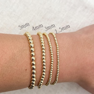 May include: Four gold beaded bracelets of varying sizes are displayed on a wrist. The bracelets are labeled 5mm, 4mm, 3mm, and 2.5mm. The bracelets are made of small, round, gold beads.