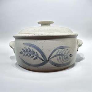 May include: A ceramic casserole dish with a lid. The dish is off-white with a speckled finish and blue floral design. It has two handles and a knob on the lid. The dish is suitable for cooking and serving.
