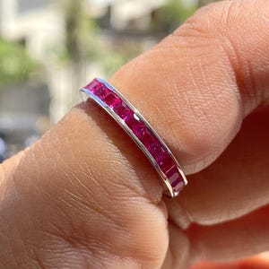 Könnte beinhalten: Ein silberner Ring mit einem Band aus quadratisch geschliffenen, leuchtend roten Edelsteinen. Der Ring ist mit mehreren Edelsteinen besetzt, wodurch eine durchgehende Farblinie entsteht. Der Ring wird an einem Finger gezeigt.