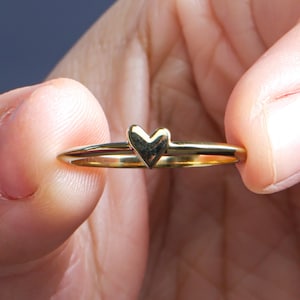 Könnte beinhalten: Ein goldfarbener Ring mit einem kleinen herzförmigen Design. Der Ring wird zwischen zwei Fingern gehalten, wodurch das zarte Band und der herzförmige Akzent hervorgehoben werden. Der Ring ist ein schlichtes, elegantes Schmuckstück.