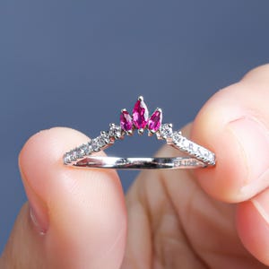 May include: A silver ring with a crown design, featuring three marquise-cut, bright pink gemstones at the centre. The band is set with small, sparkling white gemstones. The ring is held between fingers.
